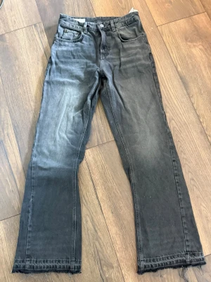 Svarta raka jeans med rå kant - Säljer ett par svarta jeans med rak passform och rå, fransad kant nedtill. Jeansen har klassisk femficksdesign och knappgylf. Materialet är denim i bomull och färgen är tvättad svart med lätt slitning. Perfekta för en avslappnad och trendig look.