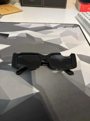 Off-White Solglasögon  - Säljer ett par svarta solglasögon med breda, rektangulära bågar och mörka glas. De har en modern och edgy look som sticker ut och passar perfekt till dig som vill ha en statement-accessoar. Glasögonen har en chunky design och är tillverkade i plast.