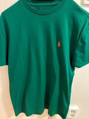 Grön t-shirt från Polo Ralph Lauren - Snygg grön t-shirt från Polo Ralph Lauren med klassisk passform och orange broderad logga på bröstet. Tillverkad i mjuk bomull och har rund halsringning samt korta ärmar. Perfekt till jeans eller shorts för en clean look.