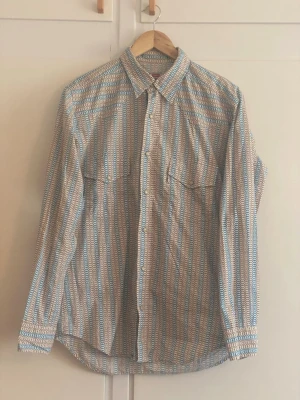Levi’s skjorta med blå ränder - Säljer en långärmad skjorta med småmönstrat tryck och vertikala blå och vita ränder. Skjortan har två bröstfickor med knapp och klassisk krage. Materialet ser ut att vara bomull, perfekt för en avslappnad men ändå stilren look.