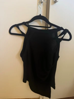Svart asymmetrisk linne med cut-outs - Snyggt svart linne med asymmetrisk nederkant och trendiga cut-outs vid axlarna. Toppen har breda axelband och är tillverkad i ett mjukt, stretchigt material som sitter skönt på kroppen. Perfekt för dig som gillar en edgy och modern stil.