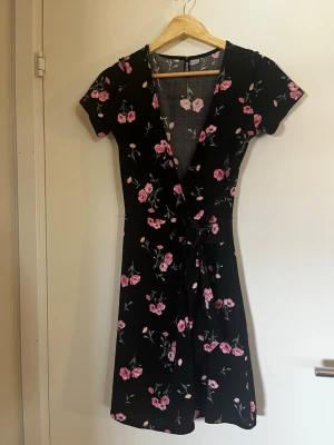 Svart omlottklänning med blommor - Svart kort omlottklänning med rosa blommigt mönster och knyt i midjan. Klänningen har v-ringning och korta ärmar, vilket ger en feminin och somrig look. Perfekt för dig som gillar romantiska detaljer och en skön passform.