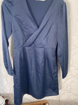 Blå omlottklänning med lång ärm - Snygg blå omlottklänning med lång ärm och v-ringad hals. Klänningen har en kort längd och är tillverkad i ett mjukt, glansigt material som ger en elegant look. Perfekt för dig som vill ha en stilren och enkel klänning med modern touch.