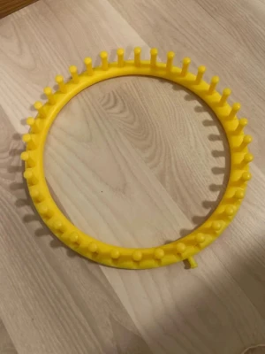 Gul stickring i plast - En rund, gul stickring i plast med piggar runt om, perfekt för att sticka mössor, halsdukar eller andra projekt utan stickor. Ringen har en diameter på cirka 29 cm och är enkel att använda för både nybörjare och erfarna stickare.