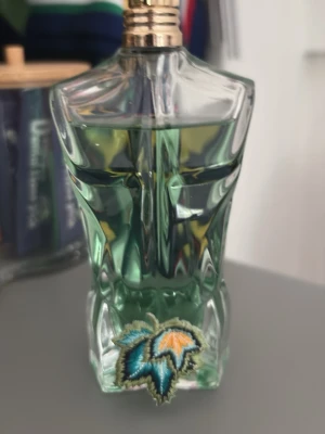 Jean Paul Gaultier Le Male parfym - Ikonisk parfymflaska från Jean Paul Gaultier formad som en manlig torso i genomskinligt glas med grön vätska. Flaskan har detaljer i guld på korken och en broderad patch med blå och orange bladmotiv framtill.