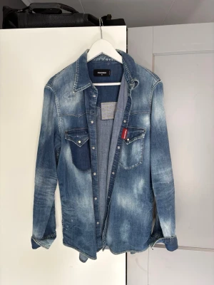 Blå jeansskjorta från Dsquared2 - Säljer en snygg blå jeansskjorta från Dsquared2 med slitna detaljer och tryckknappar. Skjortan har två bröstfickor med lock och en röd logotagg. Perfekt för dig som gillar en avslappnad men trendig stil.