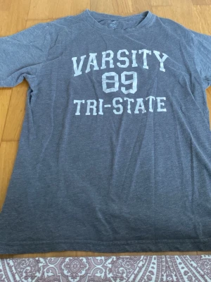 Grå t-shirt med tryck från SOC - Snygg grå t-shirt från SOC med vitt tryck på bröstet där det står 'VARSITY 89 TRI-STATE'. Klassisk rund hals och korta ärmar. Perfekt för en avslappnad stil och enkel att matcha med jeans eller shorts.