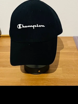 Svart Champion keps med brodyr - Snygg svart keps från Champion med klassisk böjd skärm och justerbar rem bak. Framtill sitter en vit broderad Champion-logga som ger en clean och sportig look. Tillverkad i bomullstyg som känns bekvämt och passar till de flesta outfits.