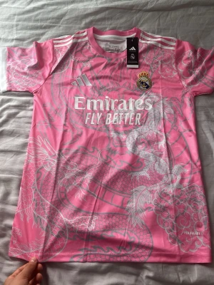 Rosa Real Madrid tröja Adidas M - Snygg rosa Real Madrid matchtröja från Adidas med drakmönster i vitt och silver. Tröjan har klubbmärke, Adidas-logga och 'Emirates Fly Better' tryckt på bröstet. Tillverkad i lätt och ventilerande material, perfekt för träning eller match. Pris kan diskuteras så lägg bud.