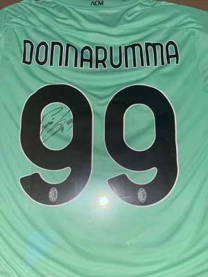 AC Milan målvaktströja signerad äkta av Donnarumma  - Säljer en mintgrön målvaktströja från AC Milan med Donnarumma och nummer 99 på ryggen. Tröjan har svarta detaljer och klubbens logga på siffrorna. Perfekt för dig som älskar fotboll och vill ha något unikt i väggen på rummet eller annatsan ram kommer med och signaturen är äkta 