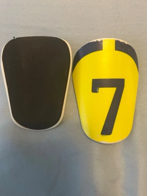 Gula benskydd med nummer 7 - Säljer ett par benskydd i sportig design, ena sidan är svart och andra sidan är gul med en stor svart siffra 7. Skydden har en böjd form för att passa smidigt mot benet och är tillverkade i plast med mjuk baksida för komfort.
