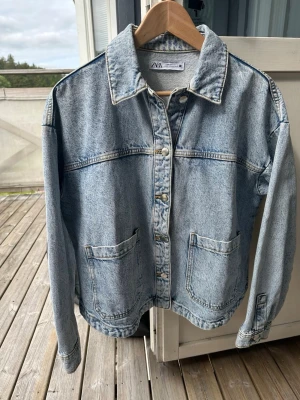Ljusblå jeansjacka från ZARA - Säljer en klassisk ljusblå jeansjacka från ZARA med raka axlar, stora framfickor och silverfärgade knappar. Jackan har en avslappnad passform och tydliga sömmar som ger en cool vintagekänsla. Perfekt att slänga över en hoodie eller t-shirt.