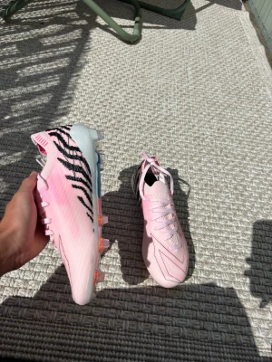 Adidas rosa fotbollsskor med zebramönster - Säljer ett par snygga Adidas fotbollsskor i ljusrosa med svarta zebraränder på hälen. Skorna har snörning, låg profil och en yttersula med färgglada dobbar för bra grepp på planen. Tillverkade i syntetmaterial och designade för fart och kontroll.