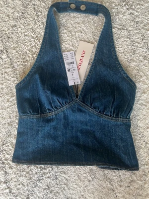 Jeans halterneck topp från Divided - Trendig jeansfärgad halterneck topp från Divided med djup v-ringning och bred resårsmock i ryggen. Toppen har tryckknappar i nacken och är croppad för en cool look. Perfekt att styla med höga jeans eller kjol.