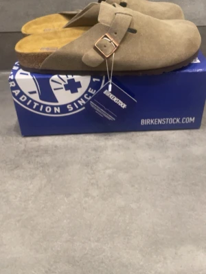 Birkenstock Boston taupe tofflor - Säljer ett par Birkenstock Boston tofflor i färgen taupe. De är tillverkade i mjukt mockaskinn med justerbart spänne i metall och har en klassisk korksula för extra komfort. Perfekta för dig som gillar stilrena och bekväma skor.