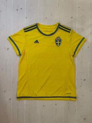 Svensk landslagströja Adidas gul - Gul svensk landslagströja från Adidas med blå detaljer och broderad landslagssköld på bröstet. Tröjan har blå ränder på axlarna, rund hals och texten 'ALTEWAISAOME' på ryggen. Tillverkad i lätt funktionsmaterial med climacool-teknologi.