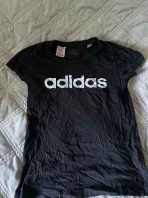 Svart t-shirt med Adidas-tryck H&M - Säljer en svart t-shirt från H&M med stort vitt Adidas-tryck på bröstet. Klassisk rund halsringning och korta ärmar. Tillverkad i mjuk bomull för en bekväm känsla. Perfekt till jeans eller shorts för en avslappnad look.