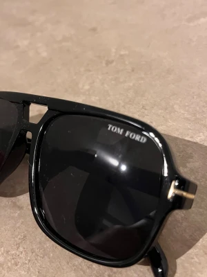 Svarta solglasögon från Tom Ford - Snygga svarta solglasögon från Tom Ford med klassisk fyrkantig form och diskreta gulddetaljer vid skalmarna. Glasögonen har en stilren design som passar perfekt till alla looks och är tillverkade i ett tåligt syntetmaterial.