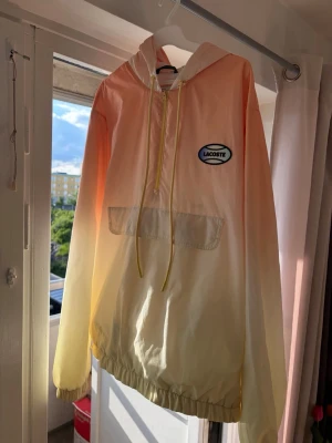 Lacoste gul-orange vindjacka med huva - Snygg vindjacka från Lacoste i gul-orange ombre med stor logga på ryggen och mindre logga på bröstet. Jackan har huva med dragsko, stor magficka och resår i ärmslut och nederkant. Perfekt för dig som vill sticka ut med en sportig och trendig look.