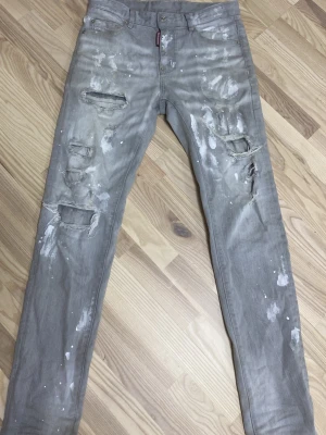 Grå slitna jeans från Dsquared2 - Säljer ett par grå jeans från Dsquared2 med distressed detaljer, hål och färgstänk för en riktigt edgy look. Jeansen har smal passform och klassisk femficksdesign. Perfekta för dig som gillar streetwear och vill sticka ut.