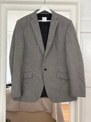 Grå kavaj från H&M med diskret mönster - Stilren grå kavaj från Bläck  med diskret rutmönster. Klassisk passform med två fickor framtill och en knappstängning. Svart innerfoder och slits bak för extra komfort. Perfekt för en dressad look.