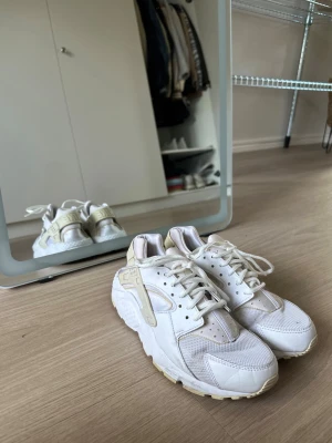 Vita Nike Huarache sneakers - Säljer ett par vita Nike Huarache sneakers med detaljer i beige. Skorna har en chunky sula, meshpaneler och klassisk Huarache-plastdetalj runt hälen. Perfekta för dig som gillar retrostil och bekväma sneakers.
