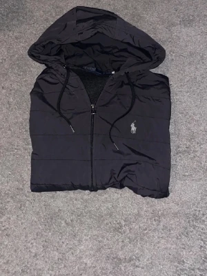 Svart pufferjacka från Polo Ralph Lauren - Svart cardigan med huva och dragkedja från Polo Ralph Lauren. Jackan har broderad logga på bröstet, dragsko i huvan och quiltad design. Materialet är glansigt och ger en sportig vibe, perfekt för kalla sommarkvällar 