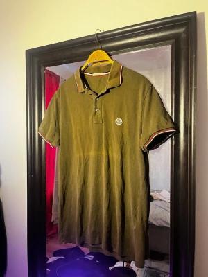 Grön Moncler pikétröja ÄKTA!!! - Säljer en grön pikétröja från Moncler med korta ärmar och klassisk krage. Tröjan har vita och röda ränder på krage och ärmslut samt Moncler-logga på bröstet. Tillverkad i bomull och har knappar vid halsen. Perfekt för en clean och stilren look. ÄKTA!!!