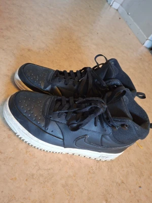 Svarta Nike high top sneakers - Snygga svarta high top sneakers från Nike med klassisk swoosh-logga på sidan. Skorna har svarta skosnören, perforerad tå och vit sula med Nike-tryck. Ovandelen är i en mix av läder och textil för en sportig look.