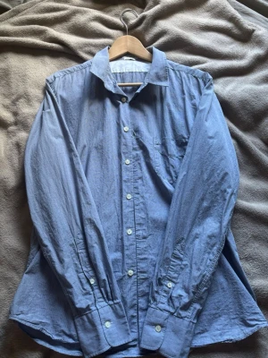 J Lindberg Blå långärmad skjorta med knappar - J Lindberg Klassisk blå skjorta med diskreta ränder, lång ärm och knäppning framtill. Perfekt till jeans eller chinos för en clean look.