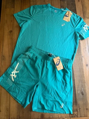 Asics Sett - Snyggt turkost träningsset från Asics med t-shirt och shorts. Setet är gjort i ett lätt och snabbtorkande material, perfekt för löpning eller gym. T-shirten har rund hals och Asics-logga på bröstet, shortsen har elastisk midja och vita detaljer på sidorna.