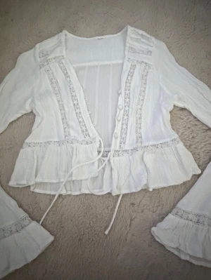 Vit boho-blus med spetsdetaljer - Romantisk vit blus i boho-stil med långa ärmar, dekorativa spetsband och knappar framtill. Blusen har volangkant nedtill och vid ärmslut samt knytband i midjan. Tillverkad i ett tunt, luftigt tyg som ger en avslappnad och trendig look.
