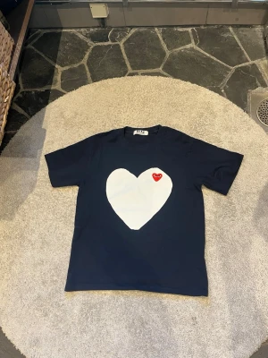 Svart t-shirt Comme des Garçons Play - Snygg svart t-shirt från Comme des Garçons Play med stort vitt hjärta och röd logga på bröstet. Klassisk passform och korta ärmar. Perfekt statement-plagg för dig som gillar streetwear och vill sticka ut.