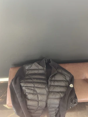 Svart Moncler cardigan - Snygg svart dunjacka från Moncler med quiltad framsida och ärmar i stickat material. Jackan har dragkedja framtill och klassisk Moncler-logga på ärmen. Perfekt för dig som vill ha en stilren och exklusiv look under kalla dagar.