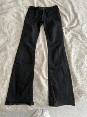 Svarta bootcut true religion  - Snygga svarta bootcut jeans med klassisk femficksdesign och knappar på bakfickorna. Jeansen har utsvängda ben och normal midja. Perfekta för dig som gillar en retroinspirerad look och vill ha ett par jeans som sticker ut lite extra. Model Becca Mid Rise Bootcut
