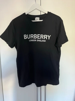 Svart Burberry t-shirt med logga - Snygg svart t-shirt från Burberry med klassisk vit logga 'BURBERRY LONDON ENGLAND' tryckt på bröstet. T-shirten har rund halsringning och korta ärmar. Tillverkad i mjuk bomull för en bekväm känsla och stilren look.