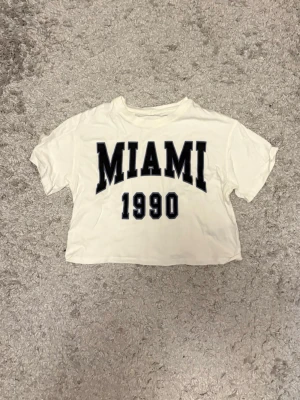 H&M Miami 1990 Cropped T-shirt Strl S  - Vit magtröja/cropped t-shirt från H&M med “MIAMI 1990”-tryck framtill. Bekväm passform och perfekt till sommaren eller för en avslappnad vardagslook.  📏 Storlek: S 🏷️ Märke: H&M ✨ Skick: Bra begagnat skick  