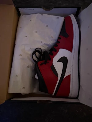 Air Jordan 1 Mid röd/svart/vit - Säljer ett par Air Jordan 1 High sneakers i klassisk röd, svart och vit färgkombination. Skorna har svart swoosh, svarta skosnören och perforerad tåbox. Tillverkade i läder med högt skaft och ikonisk Jordan-logga på plösen. Tar emot bud 