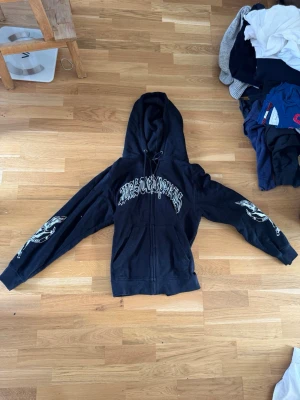 Svart hoodie från Wasted med tryck - Svart zip-hoodie från Wasted med stor vit text på bröstet och coola tryck på båda ärmarna. Hoodien har dragsko i huvan och är tillverkad i mjukt bomullsmaterial. Perfekt för dig som gillar streetwear och vill sticka ut.