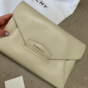  Givenchy Antigona textured leather clutch - Säljer min fina Givenchy ”Antigona textured-leather clutch”. Köptes 2018 på Net-a-Porter för 1395 amerikanska dollar, vilket motsvarar ungefär 12 000 kronor. Nästan perfekt skick, använd ett fåtal gånger. På insidan är det ett litet sträck av en bläckpenna som låg i väskan. Skriv privat för fler bilder! Tre stora fack, rymmer väldigt mycket. 2019 satte jag in två skinnhylsor med ringar hos NK skomakeri (se bild). Detta för att själv kunna sätta i kedja eller axelband och bära den som en crossbody bag. Dessa öglor är alltså inte orginal Givenchydelar, men äkta skinn. Därför går jag ner i pris. Kan tänka mig att gå ner lite lägre! Kommer med dustbag och äkthetsbevis! Äggvit färg, 30 cm bred och 21 cm hög med tre fack.  