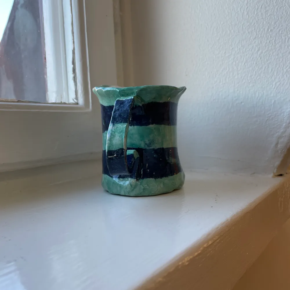 En handgjord kopp i keramik. Koppen är ljusgrön med mörkblåa ränder medan insidan av koppen är ofärgad. . Övrigt.