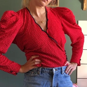 Vintage blus röd prickig puffärm - Omlottblus med puffärmar, röd med svarta prickar. I fint skick bortsett från en knapp som saknas vid ena ärmen. Inte märkt med någon storlek men uppskattas till small.