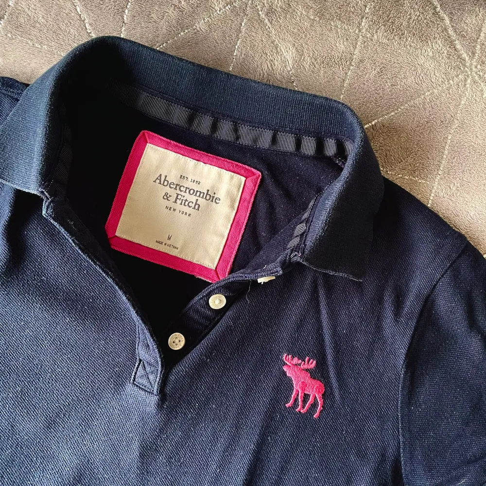 En mörkblå Abercrombie piké med rosa detaljer. I fint skick. Den är storlek M men är liten i storleken.. T-shirts.