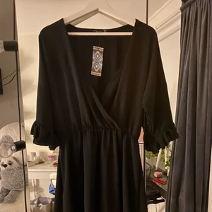 Boohoo svart omlott klänning - Svart omlott klänning från boohoo. Aldrig använt och lappen sitter på. Storlek 44 och fint skick. Klänning har en omlott urringning och längst ner på kjolen har den volanger