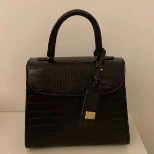 En svart handväska i krokodil skinn imitation från LYDC London inköpt på Zalando med gulddetaljer. Väskan har ett klicklås och ett innerfack med dragkedja. Ett tillhörande band medföljer samt dustbag. I toppskick då den använts knappast. Inköpt för 500 kr