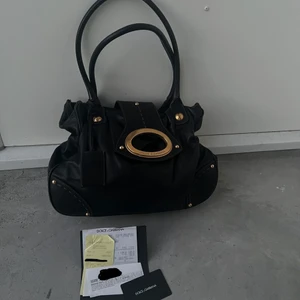 Dolce & Gabbana väska  - Vintage väska i mörk brun/svart läder från Dolce & Gabbana. Denna väska är ca 15 år gammal, köpt i LA. Röd innefoder, ett stort innerfack och ett innerfack med dragkedja. Ingen dustbag, men kvitto och authenticity card medföljer. Den är sparsamt använd, inga fläckar eller märken på lädret, endast lite spår av användning på axelremmarnas kanter, bild finns. Höjd 28, bredd 32, djup 11. Passar perfekt till bla. skolan! Skickar gärna fler bilder vid önskemål. 