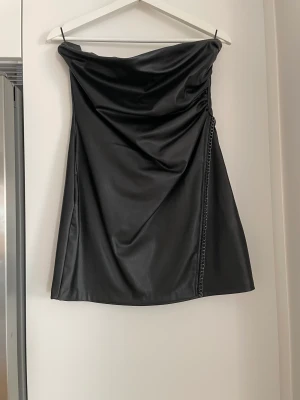 Skinnimitations klänning - En kort axelbandslös klänning i skinnimitation, med en plastad kant som fäster i stället för axelband. Cool kedjedetalj i sprunget. Kjolstycket är stängt efter sprunnet så inget syns.  Mycket gott skick, använd endast 1 gång. Stl M