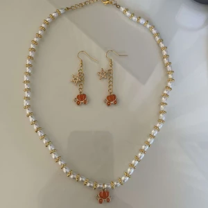 Guldhalsband och örhängen med pärlor och stjärnor - Snyggt set med ett guldhalsband och matchande örhängen. Halsbandet har vita pärlor och en orange detalj i mitten. Örhängena har stjärnor och orangea detaljer som matchar halsbandet. Perfekt för att glamma upp din outfit till fest eller speciella tillfällen!