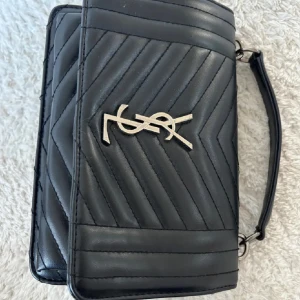 Svart quiltad axelväskafrän Yves Saint Laurent - Säljer en snygg svart axelväska frän. Väskan är quiltad med ett elegant mönster och har en guldig YSL-logga pa framsidan. Den har en kedjerem med läderdetaljer och en praktisk dragkedja inuti. Insidan är fodrad med röd textil. Perfekt för bäde vardag