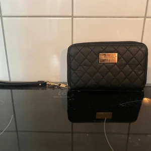 Svart quiltad plånbok - Snygg svart quiltad plånbok med dragkedja och en liten guldfärgad platta på framsidan. Perfekt för att hålla ordning på kort och kontanter. Den har en avtagbar handledsrem för enkel bärning. Passar både till vardag och fest!